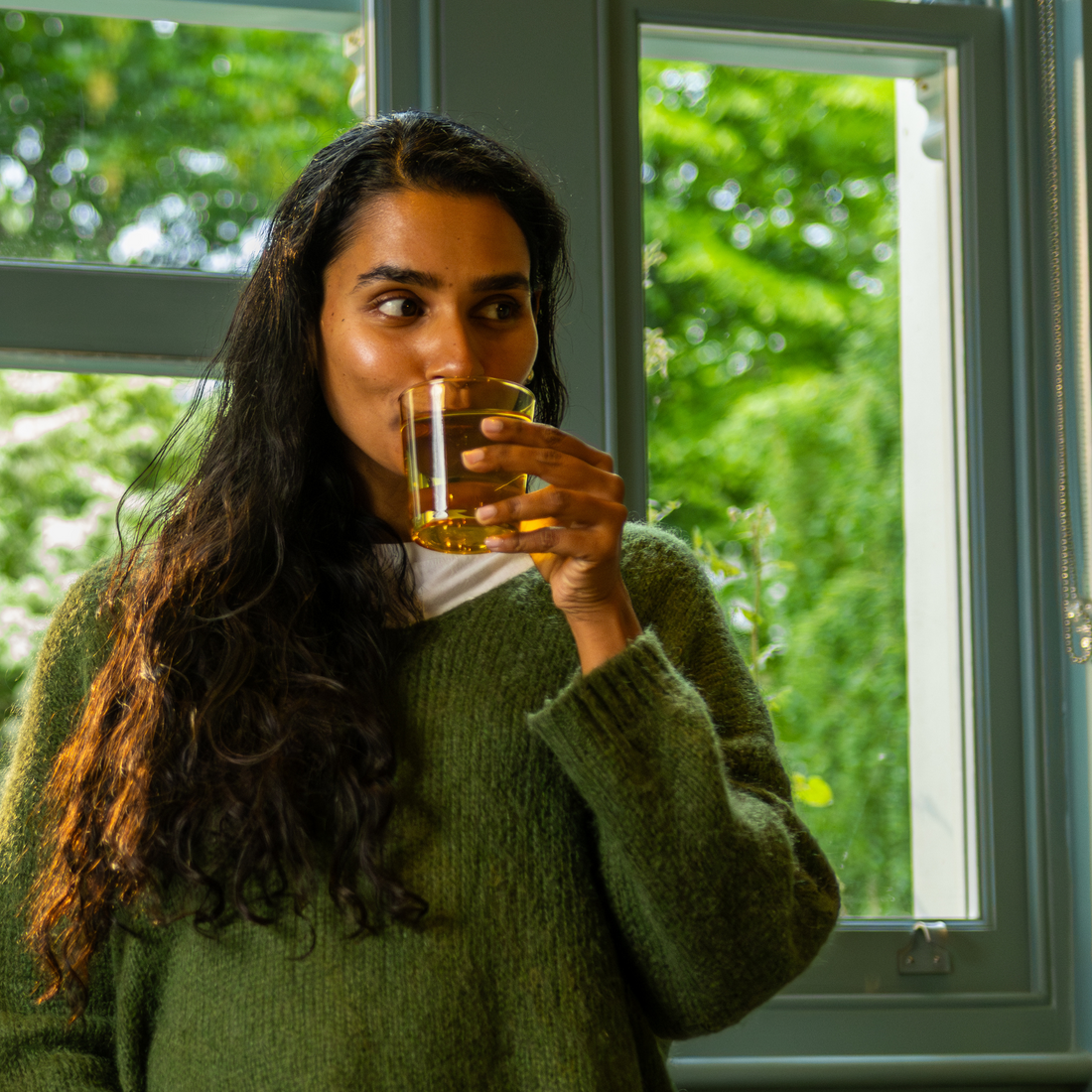 Woman in a green sweater drinking from a glass by a window with greenery outside