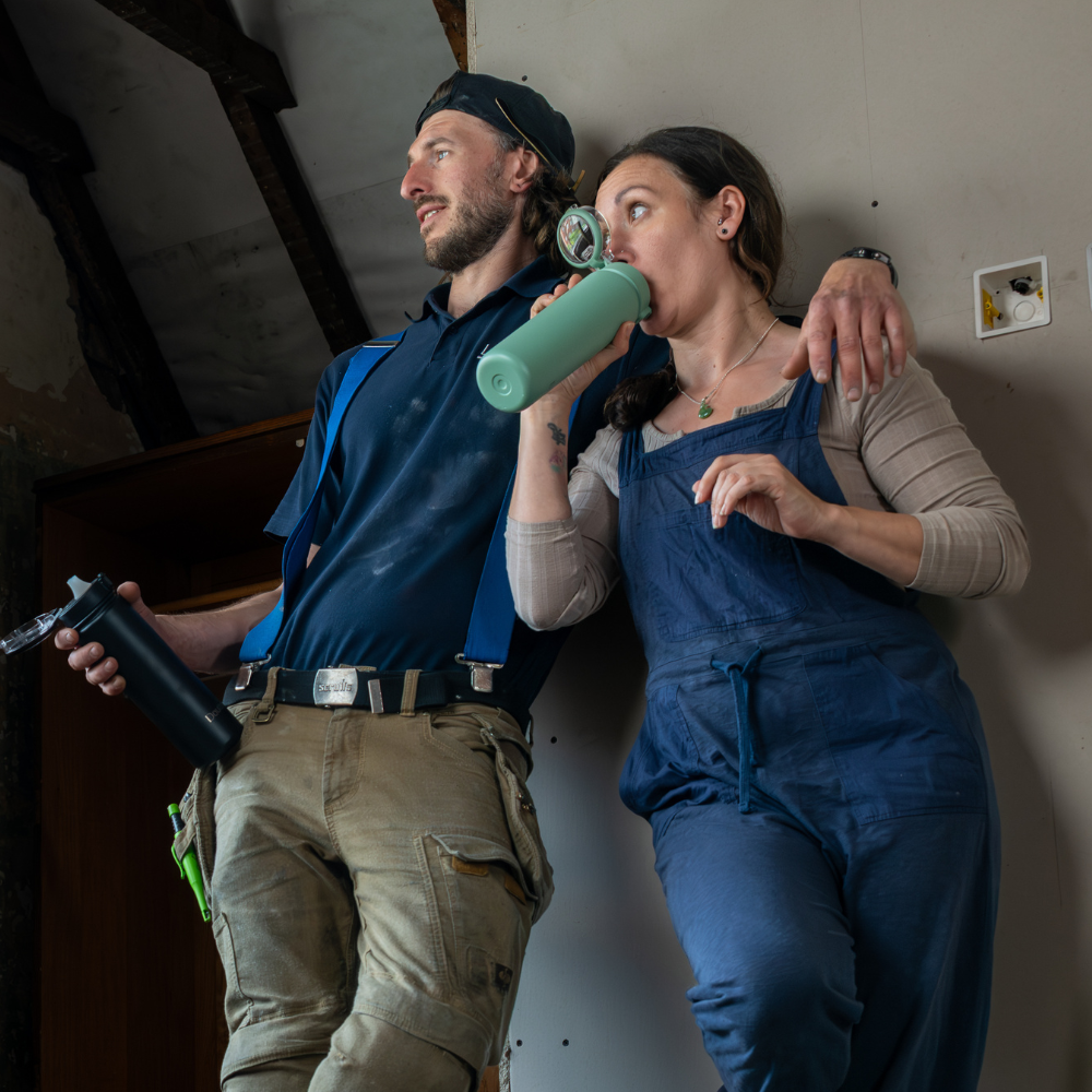 Two people in work attire, one drinking from a green bottle, against a plain wall.